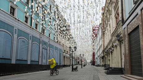 A Yandex.Eats food delivery courier rides a bicycle along a street past closed shops in Moscow