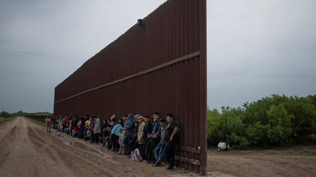 Asylum-seeking migrants from Central America line-up along the border wall as they wait to surrender to the US border patrol