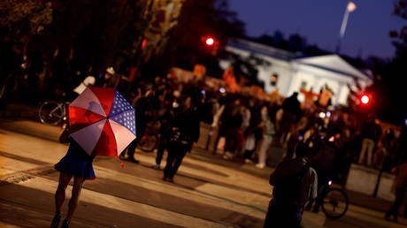 A person with an umbrella in the colors of the U.S. flag walks into a gathering of local residents waiting for the results of the 2020 presidential election outside of the White House in Washington, U.S., November 4, 2020