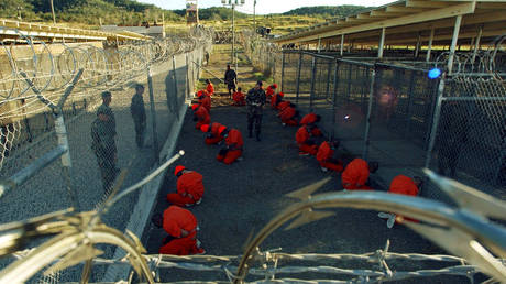 Detainees in orange jumpsuits sit in a holding area under the watchful eyes of Military Police at Camp X-Ray at Naval Base Guantanamo Bay, Cuba, during in-processing to the temporary detention facility on January 11, 2002.