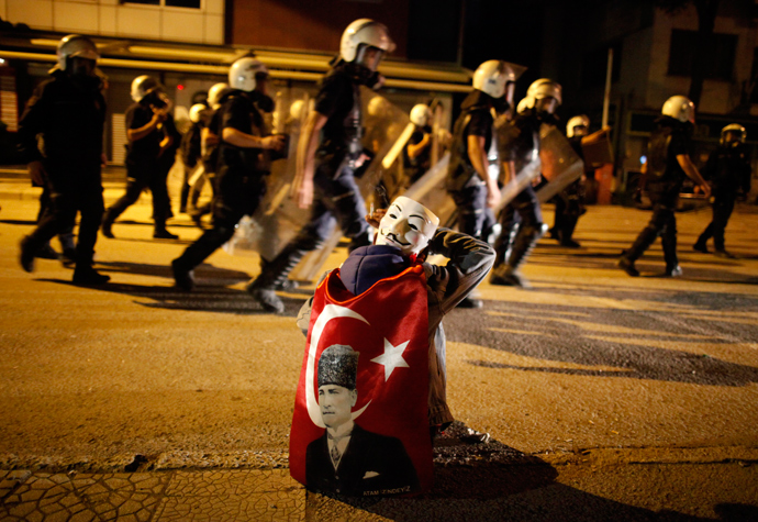 An anti-government protester with a Guy Fawkes mask behind his head smokes a cigarette as he cleans his eyes after being hit with tear gas while riot police return to their position after chasing protesters in central Ankara June 11, 2013. (Reuters / Dado Ruvic)